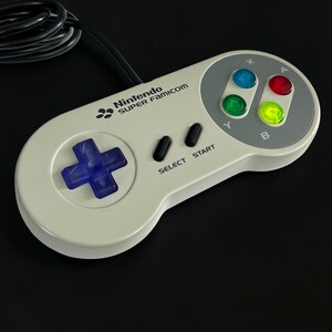 Peut inclure: Une manette de jeu vidéo Nintendo Super Famicom gris clair avec une croix directionnelle violette, deux boutons Select et Start noirs, et quatre boutons d'action translucides colorés marqués A, B, X et Y, avec un câble noir attaché.