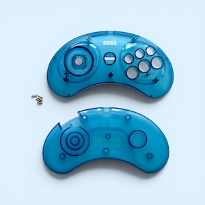 May include: Two-piece, transparent blue SEGA controller shell with the SEGA logo. The top piece has buttons and a directional pad. Small gold screws are next to the controller pieces. The controller is on a light blue background.