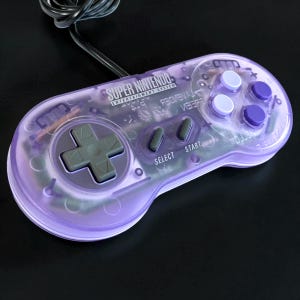 May include: A translucent purple Super Nintendo Entertainment System game controller featuring a gray directional pad, two gray center buttons labeled Select and Start, and four action buttons in light and dark purple shades with a black cord.