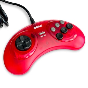 May include: A translucent red Sega video game controller featuring a black directional pad, a grey Start button, and six action buttons labeled A, B, C, X, Y, and Z in black and grey. A black wired cable extends from the top of the controller.