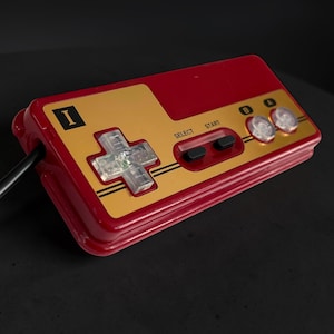 May include: A retro-style video game controller in red and gold. The controller features a directional pad, select and start buttons, and two action buttons. The controller has a black cable attached.