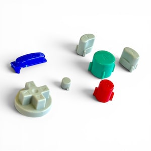 May include: A collection of colorful game controller buttons in various shapes and sizes. Includes a blue directional pad, a gray cross-shaped button, a small gray button, a green circular button, a red button, and two gray buttons.