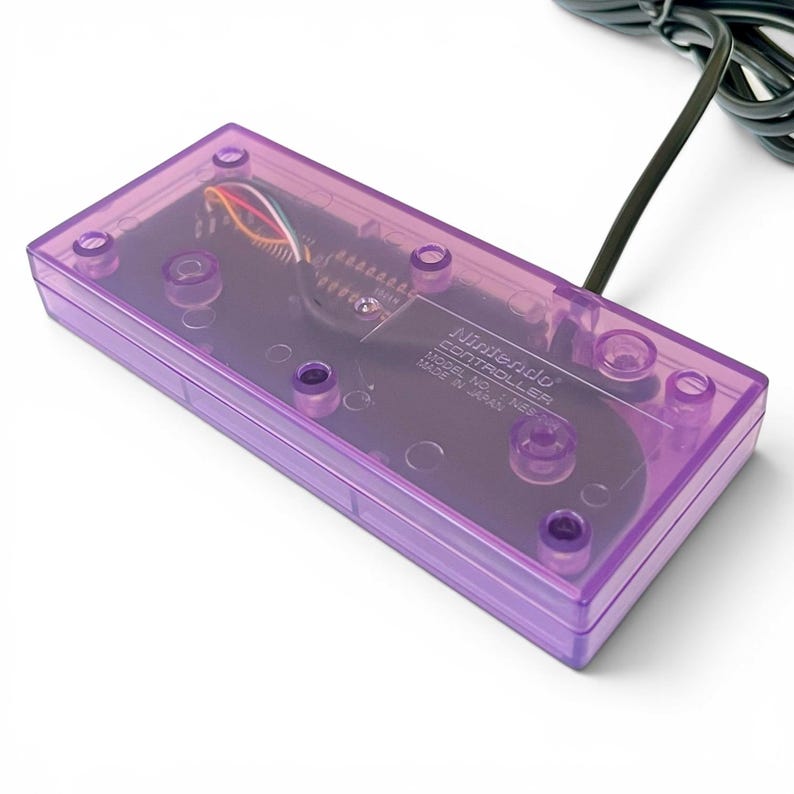 Op de afbeelding: Een transparante paarse Nintendo-controller met een zwarte kabel. De interne componenten van de controller zijn zichtbaar door de plastic behuizing. De tekst "Nintendo CONTROLLER MODEL NO. NES-004 MADE IN JAPAN" staat op de controller.