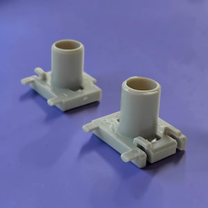 Op de afbeelding: Twee lichtgrijze kunststof onderdelen, elk voorzien van een cilindrische verticale kolom gecentreerd op een rechthoekige basis met kleine uitstekende lipjes en geïntegreerde vergrendelingsclips voor mechanische assemblage.