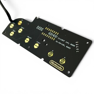 Op de afbeelding: Een zwarte Nintendo controller printplaat met goudkleurige knoplabels en tekst. De labels zijn onder meer "UP", "DOWN", "LEFT", "RIGHT", "SELECT" en "START". Het Nintendo logo is zichtbaar.
