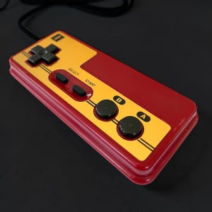 Puede incluir: Un mando de videojuegos retro rojo y dorado. El mando tiene una cruceta direccional, botones de selección e inicio, y dos botones de acción etiquetados como "A" y "B". Un cable negro se extiende desde la parte superior del mando.