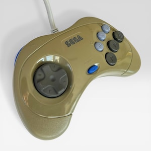 May include: A gold-colored Sega Genesis controller with a gray directional pad and buttons. The controller has a blue start button and the Sega logo is visible. The controller is connected to a white cable.