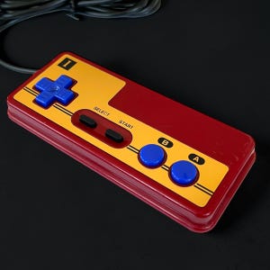 Może przedstawiać: Kontroler do gier w stylu retro w kolorze czerwono-żółtym z niebieskimi przyciskami. Kontroler posiada pad kierunkowy, dwa przyciski oznaczone jako "B" i "A" oraz przyciski "SELECT" i "START". Czarny kabel wychodzi z góry.