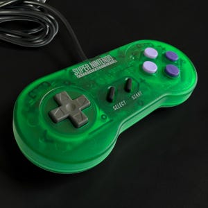 Op de afbeelding: Een doorschijnende groene Super Nintendo Entertainment System gamecontroller met een grijze richtingspad, twee zwarte select- en startknoppen en vier paarse actieknoppen. Een zwarte kabel komt uit de bovenkant van het apparaat.