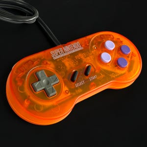 May include: A translucent orange Super Nintendo Entertainment System game controller featuring a grey directional pad, two black oval buttons labeled Select and Start, and four round action buttons in light and dark purple with a black cord.