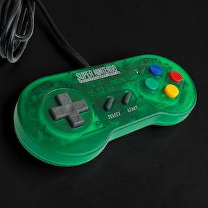 Puede incluir: Un mando de Super Nintendo verde transparente con un cable negro. El mando tiene una cruceta gris, botones de colores y el texto "SUPER NINTENDO ENTERTAINMENT SYSTEM". Los botones "SELECT" y "START" también son visibles.