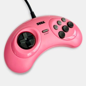 Peut inclure: Une manette de jeu vidéo Sega rose vif avec une croix directionnelle noire à gauche, un bouton Start gris au centre et six boutons d'action à droite, dont trois boutons gris marqués X, Y, Z et trois boutons noirs marqués A, B, C.