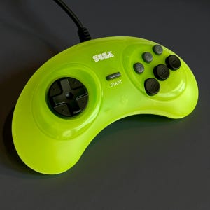 May include: A lime green Sega Genesis controller with a black directional pad, buttons, and a start button. The controller has the word "SEGA" printed on it.