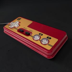 May include: A retro-style video game controller in red and gold. The controller features a directional pad, two black buttons labeled "SELECT" and "START", and two clear, round buttons labeled "B" and "A". The controller has a wired connection.