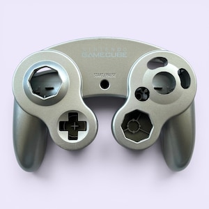 May include: A silver Nintendo GameCube controller. The controller has a D-pad, analog stick, and various buttons. The text "NINTENDO GAMECUBE" and "START/PAUSE" are visible on the controller.