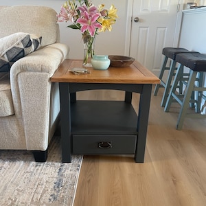 May include: A small wooden side table with a dark grey base and a natural wood top. The table has a drawer with a metal handle. A vase of pink and yellow lilies sits on top of the table, next to a small wooden bowl and a candle.