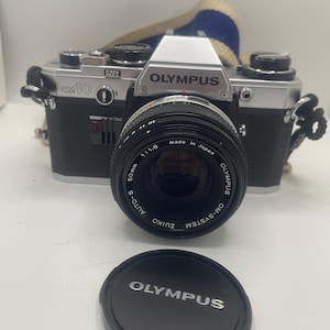以下が含まれることがあります： 50mmレンズとレンズキャップが付いた、黒とシルバーのオリンパスOM10フィルムカメラ。「ZUIKO AUTO-S 50mm 1:1.8」と表示されたレンズ。カメラには青とベージュのストラップが付いており、レンズキャップは黒で「OLYMPUS」と白で表示されています。