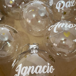 Może przedstawiać: Przezroczyste bombki choinkowe z białym napisem i złotymi akcentami. Na bombkach znajdują się słowa takie jak "Felizidad", "Paz" i "Amor". Niektóre bombki mają złote gwiazdki i serca.
