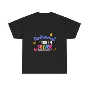 May include: A black short-sleeved t-shirt featuring a colorful graphic print that reads Professional PROBLEM SOLVER #PRINCIPALLIFE, accented with a yellow star, a pink heart, and yellow swirl doodles in blue, white, pink, and yellow text.