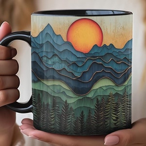 May include: A black-handled coffee mug featuring a layered mountain and forest design with a setting sun. The artwork uses shades of blue, green, and gold. The mug is held in a person's hands.