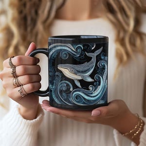 May include: Black ceramic mug featuring a detailed illustration of a whale swimming in stylized blue waves. The whale is depicted in shades of gray and white against a black background. The mug has a black handle.
