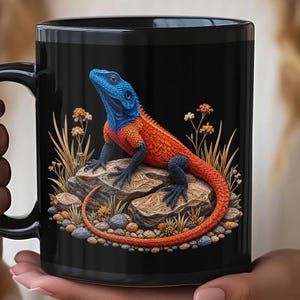 Pode incluir: Uma caneca de cerâmica preta com um gráfico vibrante de um lagarto com cabeça azul brilhante e corpo laranja. O lagarto está pousado numa rocha texturizada rodeada por pequenas pedras e caules finos de vegetação seca.