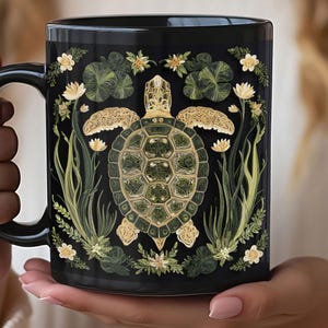 May include: Black ceramic mug featuring a detailed illustration of a sea turtle surrounded by aquatic plants and flowers. The turtle has a green and gold shell, and the design is set against a black background. The mug is held in a person's hands.