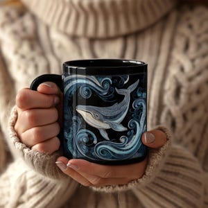 May include: Black ceramic mug featuring a detailed illustration of a whale swimming in stylized blue waves. The whale is depicted in shades of gray and white against a black background. The mug has a black handle.