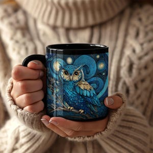 May include: A black ceramic mug featuring a detailed illustration of a blue owl perched on a branch against a swirling, starry night sky background. The owl has large, bright yellow eyes and intricate feather patterns in shades of blue and gold.