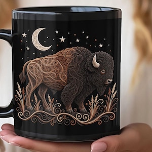 May include: A black ceramic mug featuring a detailed illustration of a bison rendered in brown and tan swirling filigree patterns. The design includes a crescent moon and stars above the bison, with wheat stalks at the base of the mug.