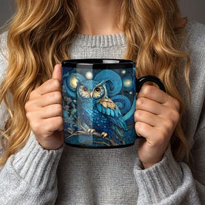 May include: A black ceramic mug featuring a vibrant blue owl perched on a branch against a swirling, starry night sky background. The owl has detailed feathers, large yellow eyes, and is surrounded by glowing golden orbs in a painterly style.