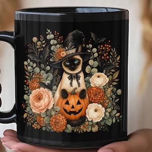 May include: A black ceramic mug featuring a Siamese cat wearing a black witch hat and a lace collar. The cat holds an orange jack-o'-lantern pumpkin, surrounded by orange and white flowers, green foliage, and small black bats on a black background.