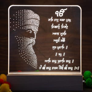 Peut inclure: Une lampe LED blanche avec une base blanche et un panneau en acrylique transparent. Le panneau présente une silhouette blanche d'une personne portant un turban et une barbe, ainsi que du texte en écriture punjabi. Le texte se lit comme suit : "96 ਸਤਿ ਨਾਮੁ ਕਰਤਾ ਪੁਰਖੁ ਨਿਰਭਉ ਨਿਰਵੈਰੁ ਅਕਾਲ ਮੂਰਤਿ ਅਜੂਨੀ ਸੈਭੰ ਗੁਰ ਪ੍ਰਸਾਦਿ ॥ ॥ ਸਾ ॥ ਆਦਿ ਸਚੁ ਜੁਗਾਦਿ ਸਚੁ ॥ ਹੈ ਭੀ ਸਚੁ ਨਾਨਕ ਹੋਸੀ ਭੀ ਸਚੁ ॥੧॥"