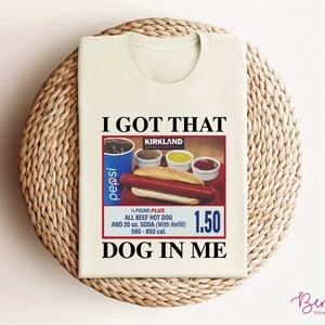 May include: A white t-shirt with the text "I GOT THAT" and "DOG IN ME" printed on it. The shirt features a vintage-style advertisement for a Kirkland Signature all-beef hot dog meal with a Pepsi, condiments, and a price tag of $1.50.