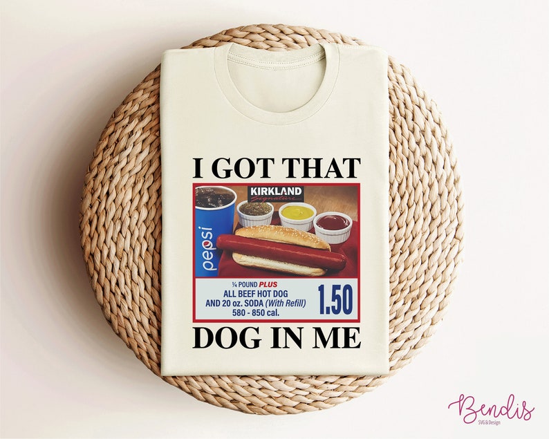 May include: A white t-shirt with the text "I GOT THAT" and "DOG IN ME" printed on it. The shirt features a vintage-style advertisement for a Kirkland Signature all-beef hot dog meal with a Pepsi, condiments, and a price tag of $1.50.