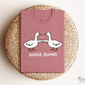 May include: A pink t-shirt with a white cartoon illustration of two geese bumping fists. The text "Goose Bumps" is printed below the geese.
