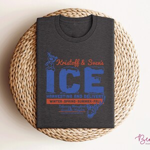 K&ouml;nnte beinhalten: Ein dunkelgraues T-Shirt mit einem blauen und roten Grafikdesign mit dem Text "Kristoff & Sven's ICE" und den Worten "Harvesting and Delivery" mit den aufgef&uuml;hrten Jahreszeiten. Die Grafik enth&auml;lt auch ein Cartoon-Bild von Olaf dem Schneemann.