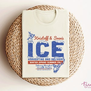 K&ouml;nnte beinhalten: Ein beiges T-Shirt mit einem Vintage-Grafikdesign, das Kristoff und Svens Eis-Ernte- und -Lieferdienst zeigt. Das Design enth&auml;lt eine blaue und wei&szlig;e Illustration von Olaf und Sven sowie den Text "Kristoff & Sven's ICE" in Blau und Rot. Der Text "Harvesting and Delivery" ist in Rot, und der Text "Winter: Spring - Summer - Fall" ist in Blau. Der Text "Delivering throughout Arendelle Kingdom" ist in Blau, und der Text "Contact Sven for details" ist in Rot.