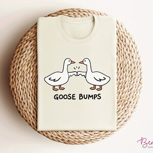 May include: A light beige t-shirt with a graphic of two cartoon geese bumping fists. The text "GOOSE BUMPS" is printed below the geese.