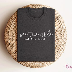 K&ouml;nnte beinhalten: Ein dunkelgraues T-Shirt mit dem Text "see the able not the label" in wei&szlig;en Buchstaben.