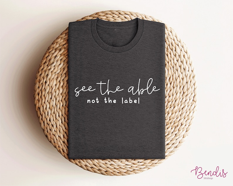 K&ouml;nnte beinhalten: Ein dunkelgraues T-Shirt mit dem Text "see the able not the label" in wei&szlig;en Buchstaben.
