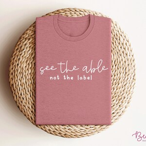 K&ouml;nnte beinhalten: Ein pinkfarbenes T-Shirt mit dem Text "see the able not the label" in wei&szlig;en Buchstaben.