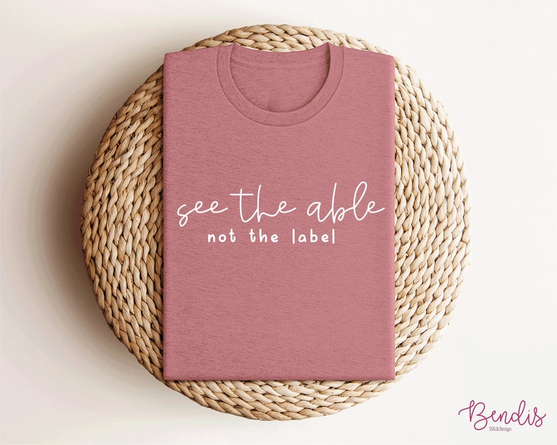 K&ouml;nnte beinhalten: Ein pinkfarbenes T-Shirt mit dem Text "see the able not the label" in wei&szlig;en Buchstaben.