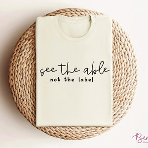 K&ouml;nnte beinhalten: Ein beiges T-Shirt mit dem Text "see the able not the label" in Schwarz gedruckt.