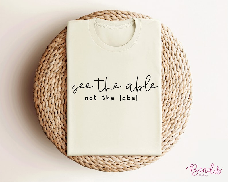 K&ouml;nnte beinhalten: Ein beiges T-Shirt mit dem Text "see the able not the label" in Schwarz gedruckt.