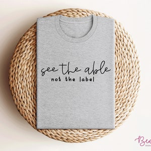 K&ouml;nnte beinhalten: Graues T-Shirt mit dem Text "see the able not the label" in schwarzer Schrift.