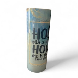 May include: A tall, cylindrical tumbler with a blue and gold marbled design. The tumbler features the words "HOLD with a high HOO she is strong PROVERBS" in a bold, stylized font. The overall aesthetic is modern and inspirational.