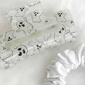 Op de afbeelding: Een doorzichtige haarclip met een wit spookpatroon en een witte satijnen scrunchie met een zwart label. De scrunchie heeft een zwart label. De items liggen op een wit, getextureerd oppervlak.