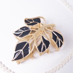 May include: A gold-toned metal hair claw clip in a leaf design with black enamel detailing. A strand of white faux pearls curves across the white background, adding a touch of elegance to the hair accessory.