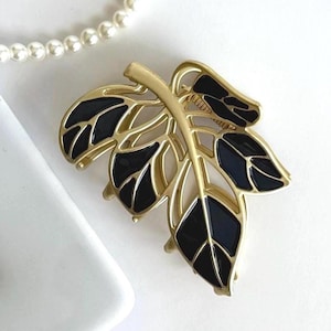 May include: Gold-toned leaf brooch with black enamel detailing. The brooch features a detailed leaf design with a gold stem and veins. A white pearl necklace is in the background. Jewelry.
